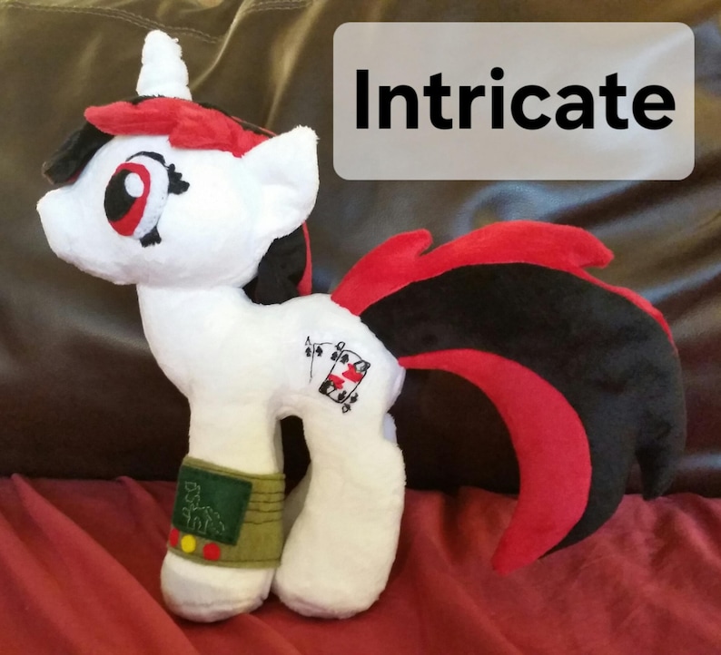 Pode incluir: Unic&oacute;rnio de pel&uacute;cia branco com crina e cauda vermelhas e pretas. O unic&oacute;rnio tem um olho vermelho, um nariz preto e uma boca preta. O unic&oacute;rnio tem um s&iacute;mbolo de naipe de cartas preto e vermelho no seu lado. O unic&oacute;rnio est&aacute; usando uma pulseira verde e amarela com o texto "D.S.F.N.L." nela.