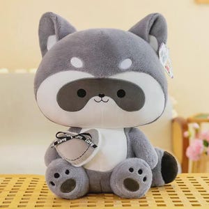 May include: A plush raccoon toy with soft gray and white fur, featuring a dark gray eye mask, small ears, and a heart-shaped pillow held in its arms. The pillow is light gray with a black ribbon printed with the word LOVE.