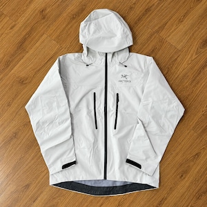 Può includere: Una giacca con cappuccio grigio chiaro dotata di una cerniera frontale nera a tutta lunghezza, due tasche verticali sul petto con cerniera nera e maniche lunghe con polsini neri regolabili. Il logo Arc'teryx è stampato in nero sul petto a sinistra.
