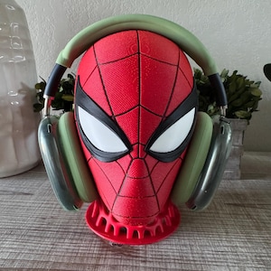 Puede incluir: Un soporte de exhibición con forma de cabeza de Spider-Man en rojo y negro, con superficie texturizada y ojos blancos. El soporte lleva puestos unos auriculares con diadema y almohadillas de color verde salvia y copas gris metálico.