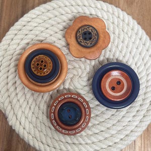 May include: Four circular buttons with varied designs. One is a light brown flower shape with a dark center. Another is a large light brown button with a dark blue center. A third is a dark blue button with a light brown center. The last is a brown button with white stitching details and a dark blue center.