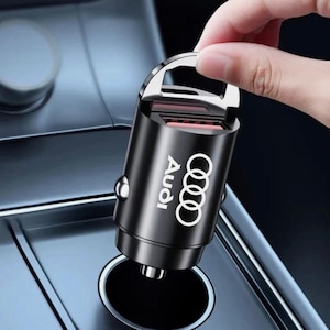May include: A black cylindrical car charger featuring the white Audi logo and four interlocking rings printed on the side. The device has a metallic finish, a top handle loop, and a standard plug connector at the base for a vehicle power outlet.