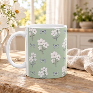 Könnte beinhalten: Ein weißer Keramikbecher mit einem wiederkehrenden Muster aus kleinen weißen Blumensträußen und hellblauen Schleifen auf salbeigrüner Basis. Der Becher hat einen Standardhenkel und eine zylindrische Form mit floralem Design.