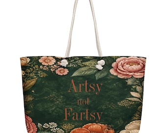 May include: A dark green tote bag featuring a floral pattern with pink and orange flowers and the text Artsy not Fartsy printed in the center. The bag has two white rope handles attached to the top with silver-toned metal grommets.