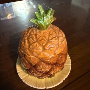 May include: A ceramic pineapple-shaped lidded jar with a textured golden-brown body and a green leafy top, resting on a round, pale yellow scalloped plate. The surface of the jar features a detailed, raised diamond pattern mimicking pineapple skin.