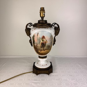 May include: A white porcelain table lamp with a hand-painted oval scene of a person holding a child. The lamp features ornate bronze-toned metal handles shaped like lion heads, a decorative bronze base, and a brass-colored light socket fixture.