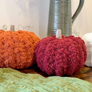 May include: Three handmade pumpkins crafted from textured, fuzzy yarn in shades of burnt orange, deep burgundy, and white. Each pumpkin features a small, light-colored wooden stem at the top and thin, curled wire tendrils extending outward.