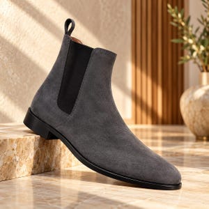 Op de afbeelding: Een antracietgrijze suède Chelsea boot met een zwart elastisch zijpaneel, een bijpassende zwarte treklus bij de hiel en een gladde zwarte zool. De enkelhoge laars heeft een ronde neus en een strak, gestroomlijnd silhouet met zichtbare stiksels.
