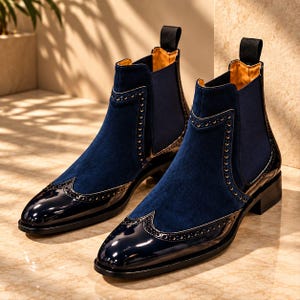 Op de afbeelding: Een paar marineblauwe Chelsea-laarzen met een combinatie van suède en glanzend lakleer. De laarzen hebben een wingtip-neus met decoratieve brogue-perforaties, elastische zijpanelen, een zwarte treklip en een lage gestapelde hak.