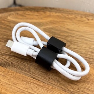Pode incluir: Um cabo de carregamento USB-C branco enrolado em um laço e preso com dois clipes de organização de cabos retangulares pretos. O cabo possui um conector USB-C padrão em uma extremidade, com o fio branco apresentando um acabamento levemente texturizado.