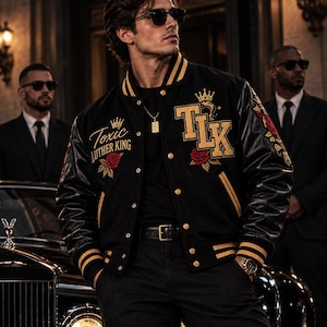 May include: A black varsity jacket with gold striped ribbed trim, featuring embroidered red roses, crowns, and the text "Toxic LUTHER KING" and "TLK" in gold lettering. The jacket has leather sleeves, gold snap buttons, and side pockets.