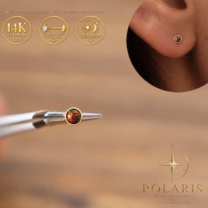 Könnte beinhalten: Ein 14-karätiger Goldstecker mit einem runden, dunklen, schillernden Opal. Das Schmuckstück wird mit einer Metallpinzette gehalten und an einem Ohrläppchen getragen. Text enthält 14K GOLD, THREADLESS PUSH PIN, 24/7 WEAR und POLARIS.