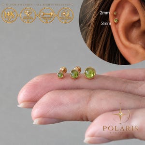 May include: Three 14K gold flat back stud earrings featuring round green gemstones. The studs are shown in 2mm and 3mm sizes, with one earring inserted into an ear cartilage piercing. The text Polaris Fine Jewelry is visible.