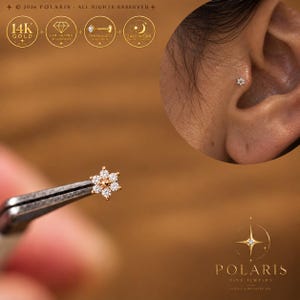 May include: A 14K gold flower-shaped stud earring featuring six small lab-grown diamonds surrounding a central gold bead. The jewelry is a threadless push-pin design suitable for 24/7 wear, shown held by tweezers and inserted into a tragus ear piercing.