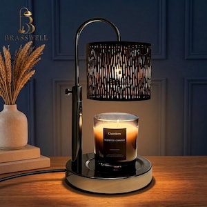 May include: A black metal candle warmer lamp with a cylindrical shade featuring vertical cutouts. The lamp base holds a glass jar candle labeled Clairière Scented Candle. The lamp has a curved arm and a circular base with a power cord.