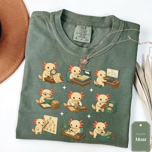 May include: A moss green crew neck t-shirt featuring a graphic print of nine light pink axolotls engaged in various office tasks, including reading, using a computer, and filing documents. The shirt has a Comfort Colors brand label at the neck.