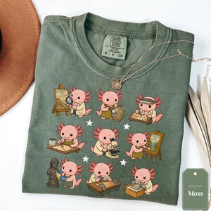 May include: A moss green t-shirt featuring a graphic print of nine pink axolotls engaged in various artistic and scientific activities, including painting, sculpting, and using a microscope. The shirt has a Comfort Colors brand label.