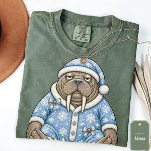 May include: A moss green t-shirt featuring a graphic of a brown walrus wearing a blue pajama set with white snowflake patterns, a matching nightcap, and silver bows. The shirt has a Comfort Colors label and a tag labeled Moss.