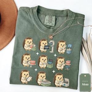 May include: A moss green crew neck t-shirt featuring a graphic print of nine hedgehogs dressed as dentists performing various dental procedures, including examining teeth, using X-ray machines, and holding dental tools and models.