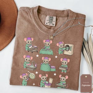 Könnte beinhalten: Ein braunes Kurzarm-T-Shirt mit einem Grafikdruck von neun Zeichentrickmäusen, die in OP-Kleidung und lila Hauben gekleidet sind. Die Mäuse sind bei verschiedenen medizinischen Aufgaben dargestellt, wie Händewaschen und der Nutzung von Instrumenten.