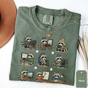 Op de afbeelding: Een mosgroen t-shirt met ronde hals en een grafische print van negen cartoon wasberen die verschillende kantoor- en gameactiviteiten uitvoeren, waaronder het gebruik van computers, headsets en grafieken, met een Comfort Colors-label.