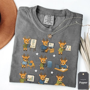 Könnte beinhalten: Ein anthrazitfarbenes T-Shirt mit einem wiederkehrenden Muster aus Zeichentrick-Rehen, die verschiedene Büro- und Datenanalyseaufgaben ausführen, darunter die Arbeit an Computern, Diagrammen und Lupen, durchsetzt mit kleinen weißen Sternen.