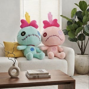 May include: Two plush dolls with large round heads, stitched mouths, and button eyes. The left doll is mint green with a yellow hand-shaped crest, while the right doll is light pink with a pink hand-shaped crest. Both feature bright pink bows.