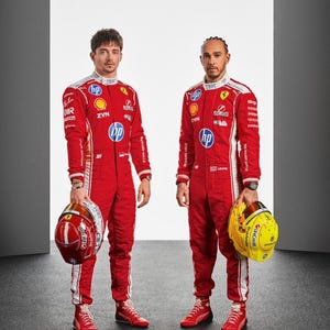 May include: Two race car drivers wearing matching red and white racing suits with various sponsor logos including HP, Shell, and ZYN. One driver holds a red and white helmet, while the other holds a bright yellow helmet with black accents.
