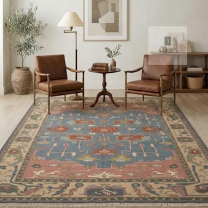 May include: A large rectangular area rug featuring a central blue field with intricate floral and geometric patterns in red, green, and cream. The rug has a wide cream-colored border with repeating motifs and a short fringe along the edges.