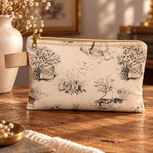 Op de afbeelding: Een rechthoekig etui met rits van gebroken wit textiel met een zwarte inktprint van bomen, stadsgebouwen, vogels en figuren in diverse houdingen. Het etui is voorzien van een goudkleurige metalen rits en een beige stoffen lus aan de zijkant.