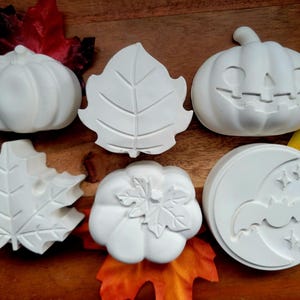 May include: A collection of six white, unpainted plaster figures featuring two pumpkins, one jack-o'-lantern, two maple leaves, and a circular disc with a bat, crescent moon, and stars, arranged alongside several colourful artificial autumn leaves.
