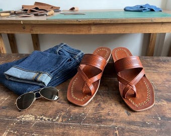 May include: A pair of brown leather sandals with a toe loop, embossed strap detailing, and gold-tone metal eyelets along the edges, accompanied by a pair of dark blue denim jeans and gold-rimmed aviator sunglasses with dark lenses.