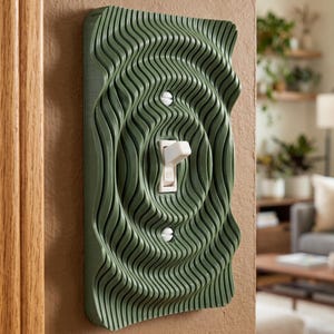 May include: A sage green light switch cover plate featuring a three-dimensional rippled, wave-like texture. The rectangular cover is mounted on a wall and surrounds a standard white toggle light switch, secured by two visible white screws.