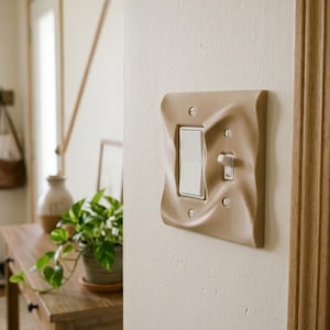 May include: A beige light switch wall plate with a wavy, rippled texture. The rectangular cover features a large white rocker switch on the left and a smaller white toggle switch on the right, secured to the wall with four silver screws.
