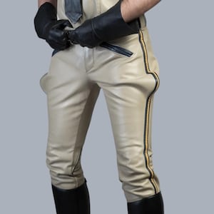 May include: Beige leather trousers featuring black leather side stripes with thin yellow piping, a black leather tie, and black leather gloves. The trousers have a front zip closure and are paired with black leather knee-high boots.