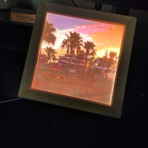 Può includere: Una cornice quadrata illuminata che mostra una scena tropicale retroilluminata con palme e un cielo al tramonto nei toni dell'arancione, rosa e giallo. La cornice ha un bordo in legno chiaro ed emette una luce calda da dietro l'immagine.