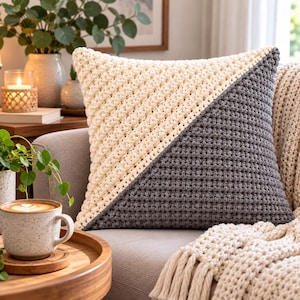 May include: A square throw pillow featuring a diagonal color-blocked design. One half of the pillow is crafted from cream-colored yarn, while the other half is made from dark gray yarn, both showcasing a textured, crocheted stitch pattern.