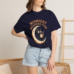 Könnte beinhalten: Ein kurzärmeliges T-Shirt in Marineblau mit einer Grafik eines schwarzen Katzenwesens, das eine Brille trägt und ein Buch liest, während es auf einer Mondsichel sitzt. Der Text "MIDNIGHT READERS CLUB" ist in einem Bogen hellorange aufgedruckt.