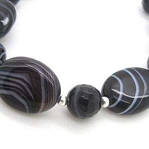 Puede incluir: Un brazalete de &aacute;gata negra y blanca con cuentas de forma ovalada. Las cuentas tienen un patr&oacute;n rayado y est&aacute;n separadas por separadores plateados.