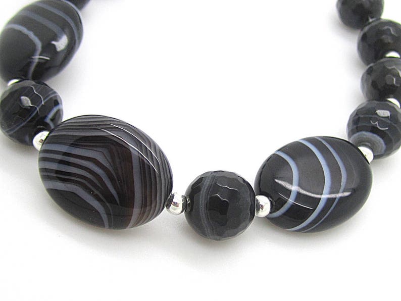Puede incluir: Un brazalete de &aacute;gata negra y blanca con cuentas de forma ovalada. Las cuentas tienen un patr&oacute;n rayado y est&aacute;n separadas por separadores plateados.