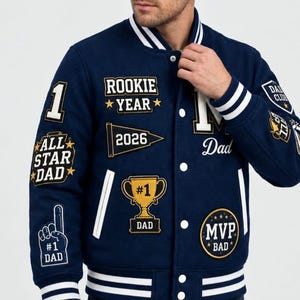May include: A navy blue varsity jacket featuring white striped ribbed trim and various embroidered patches including the text Rookie Year, 2026, All Star Dad, #1 Dad trophy, MVP Dad, and Dad Club. The jacket has white snap buttons.