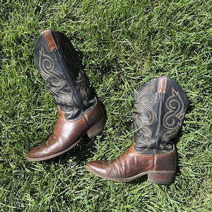 Op de afbeelding: Een paar tweekleurige cowboylaarzen met donkerbruine leren voorvoeten en zwarte leren schachten. De schachten zijn versierd met ingewikkeld beige stiksel in een wervelend patroon. Elke laars heeft een puntige neus, een hak en aantreklussen.