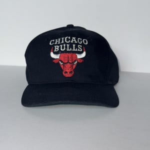 Puede incluir: Una gorra de béisbol negra con el logotipo de los Chicago Bulls bordado en la parte delantera. El diseño incluye el texto CHICAGO BULLS en letras blancas sobre un gráfico de cabeza de toro rojo y blanco con contornos negros, centrado.