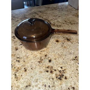 May include: A translucent brown glass saucepan with a matching circular lid and a long, integrated handle. The cookware features a smooth, reflective surface and a deep, rounded basin, showcasing a uniform amber-tinted glass construction.
