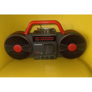 May include: A Magnavox Roller portable radio and cassette player featuring a dark grey body with a bright red handle and speaker accents. The front displays the Magnavox logo, the word roller, a radio tuner, a cassette deck, and control knobs.