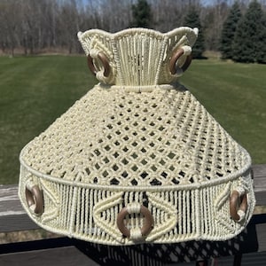 May include: A cream-colored macrame lampshade featuring a complex diamond-patterned knot design. The shade is accented with circular wooden rings integrated into the top and bottom borders, creating a textured, woven appearance throughout.