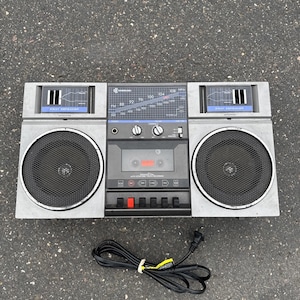 May include: A vintage Samsung portable radio cassette player in a grey plastic housing with two circular black mesh speakers, a central analogue radio tuner dial, a cassette deck, and a black power cord with a two-pin plug attached at the bottom.