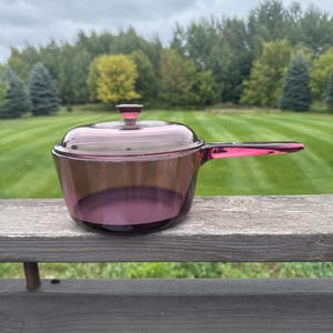 Pode incluir: Uma panela de vidro roxo translúcido com tampa a condizer e uma pega longa integrada. O utensílio de cozinha apresenta um corpo liso e arredondado com um botão circular no centro da tampa, exibindo um tom violeta profundo no vidro.