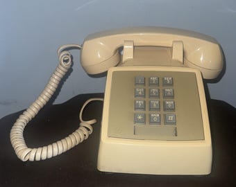 May include: A beige landline telephone with a rectangular base and a matching handset resting on top. The device features a central keypad with grey buttons, each marked with numbers, letters, and symbols, connected by a coiled cord.