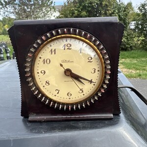 May include: A dark brown, square-shaped Telechron electric clock with a circular face, cream-colored dial, and black numerals. The clock features a metallic gold-toned bezel with a gear-like edge, black hands, and the text Telechron and Selector.
